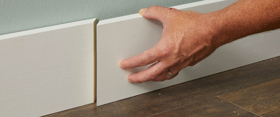 Close-up of a person installing a white baseboard along a wall, aligning two pieces for a seamless joint. The baseboard is set against a green wall and dark wood flooring, demonstrating a precise installation process.