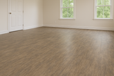 Sunlit room featuring Wilsonart laminate flooring in warm brown tones. The room has light beige walls, white baseboards, and two large windows letting in natural light, creating soft reflections on the textured wood-look laminate planks. 